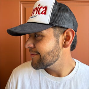 K&ouml;nnte beinhalten: Schwarz-wei&szlig;e Trucker-M&uuml;tze mit dem Wort "America" in roten Blockbuchstaben auf der Vorderseite. Die M&uuml;tze hat eine wei&szlig;e Frontplatte und eine schwarze Mesh-R&uuml;ckseite. Der Schirm ist schwarz.