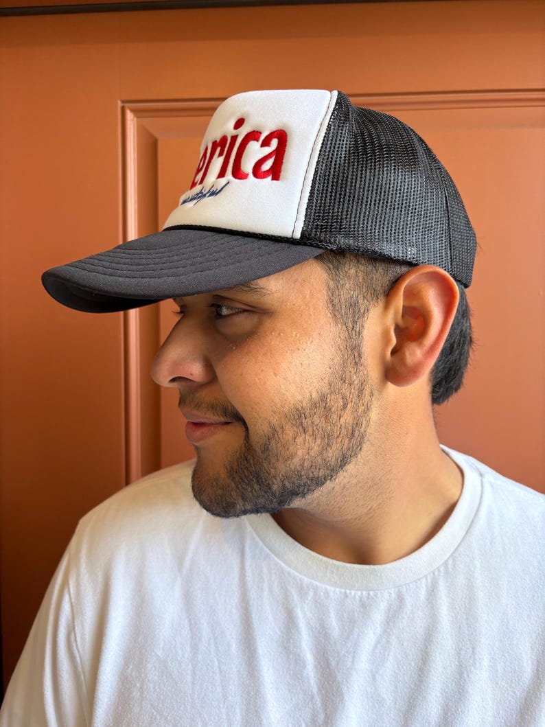 K&ouml;nnte beinhalten: Schwarz-wei&szlig;e Trucker-M&uuml;tze mit dem Wort "America" in roten Blockbuchstaben auf der Vorderseite. Die M&uuml;tze hat eine wei&szlig;e Frontplatte und eine schwarze Mesh-R&uuml;ckseite. Der Schirm ist schwarz.