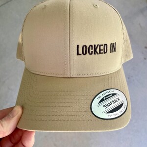 Peut inclure: Casquette snapback beige avec l'inscription "LOCKED IN" brodée en noir. La casquette a une visière incurvée et un dos en filet. Un autocollant "Yupoong Snapback" blanc et noir est sur la visière.