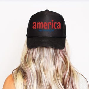 K&ouml;nnte beinhalten: Schwarze Trucker-M&uuml;tze mit dem Wort "America" in roten Buchstaben und "the beautiful" in blauer Schrift. Die M&uuml;tze wird von einer Person mit blondem Haar getragen.