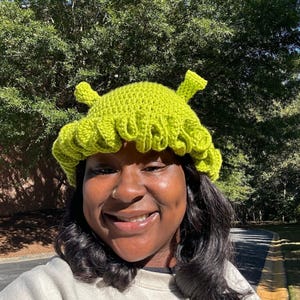 May include: A lime green crocheted hat with a ruffled brim and two small, upright protrusions. The hat is a vibrant shade of green and appears to be handmade. The hat is a whimsical accessory.