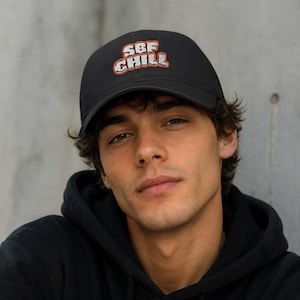 May include: A black baseball cap featuring the embroidered text SBF CHILL in white letters with a bold orange outline. The cap has a structured crown and a curved brim, constructed from a solid black fabric material with matching stitching.