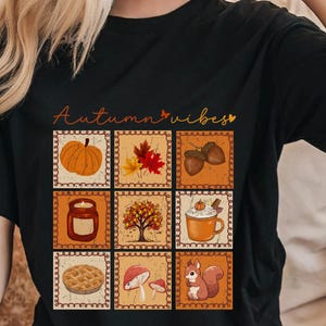 May include: A black t-shirt featuring the text Autumn vibes in orange script. Below the text is a grid of nine square illustrations depicting a pumpkin, autumn leaves, acorns, a candle, a tree, a mug, a pie, mushrooms, and a squirrel.