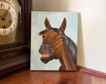 May include: A rectangular canvas painting featuring a brown horse with a black mane and forelock, shown with its mouth slightly open to reveal teeth. The horse is set against a light blue, brush-stroked background, with a small signature in the corner.