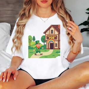 Puede incluir: Camiseta blanca con un gráfico colorido de un duende caminando hacia un pub con un arcoíris. El letrero del pub dice "POT OF GOLD PUB". El diseño tiene acentos verdes, marrones y amarillos, ideal para el Día de San Patricio.