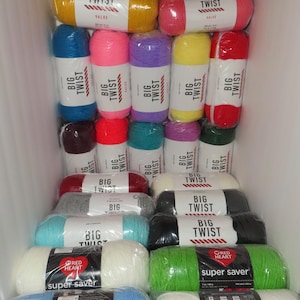 May include: A variety of colorful yarn skeins, including blue, yellow, pink, and green, are stacked in a white storage unit. The yarn is labeled "Big Twist" and "Super Saver" with the "Red Heart" logo. The image showcases a selection of craft supplies.