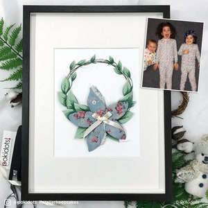 Pode incluir: Obra de arte emoldurada com um design floral em azul e rosa, rodeada por uma coroa de folhas verdes. Inclui uma pequena foto de três crianças. A obra de arte está numa moldura preta.