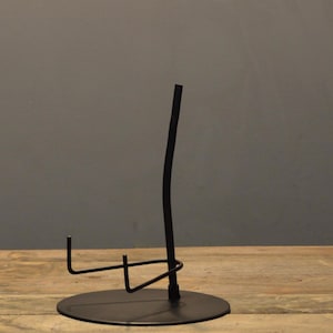 May include: A black metal display stand with a circular base and a curved, vertical support. The stand features a horizontal arm and a small hook, likely for holding an item. The design is minimalist and modern, suitable for showcasing various objects.