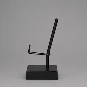 Puede incluir: Un soporte de exhibición negro mate con una base rectangular y una varilla de soporte vertical que cuenta con un brazo de apoyo en forma de U inclinado. La estructura metálica está diseñada para sostener objetos en posición vertical.
