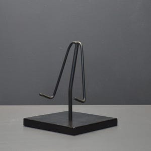 Op de afbeelding: Een zwarte metalen displaystandaard met een vierkante basis en een gebogen bovenkant. De standaard is ontworpen om een klein object vast te houden, zoals een bord of een ingelijste foto. De standaard is gemaakt van metaal en heeft een matte afwerking.