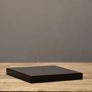 Könnte beinhalten: Ein quadratischer, schwarzer Holz-Präsentationsständer. Der Ständer hat eine einfarbige, dunkle Farbe und ein schlichtes, minimalistisches Design. Er steht auf einer Holzoberfläche mit einem neutralen grauen Hintergrund.