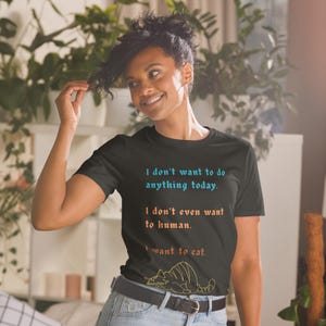 Puede incluir: Camiseta negra con el texto "I don't want to do anything today. I don't even want to human. I want to cat." en azul y naranja. Una ilustración de gato amarillo en la parte inferior.