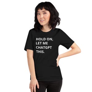 Könnte beinhalten: Schwarzes T-Shirt mit dem Text "HOLD ON, LET ME CHATGPT THIS." Das Shirt hat kurze Ärmel und einen Rundhalsausschnitt. Die Person trägt eine schwarze Hose.