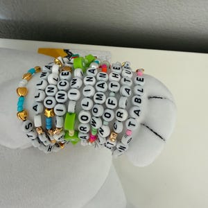 May include: A collection of beaded bracelets with white letter beads spelling out phrases like "It's all ways on", "Carolina Woman", and "Incredible". The bracelets feature gold, turquoise, and pink accents.