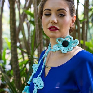 Pu&ograve; includere: Una donna indossa un vestito blu con una collana di fiori all'uncinetto. La collana &egrave; blu chiaro con un centro nero e ha dei pon pon.