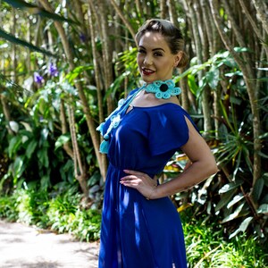 Pu&ograve; includere: Una donna con un vestito blu e una collana di fiori all'uncinetto. La collana &egrave; blu turchese con centri neri sui fiori. La donna &egrave; in piedi in un ambiente da giardino.