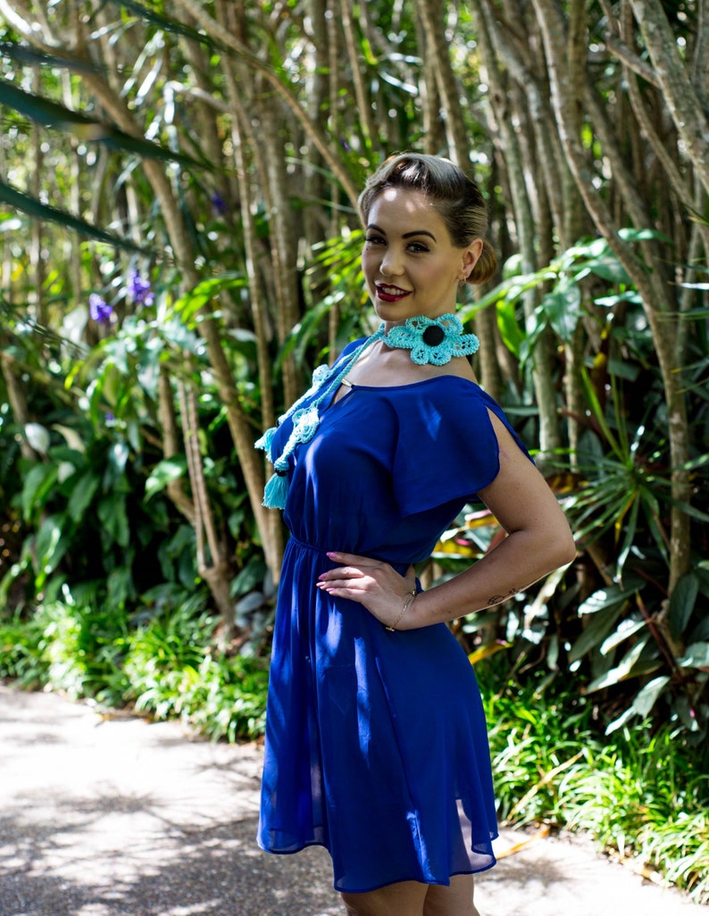 Pu&ograve; includere: Una donna con un vestito blu e una collana di fiori all'uncinetto. La collana &egrave; blu turchese con centri neri sui fiori. La donna &egrave; in piedi in un ambiente da giardino.