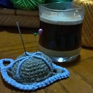 Può includere: Un cuscino per spilli in crochet blu con un centro grigio e tre spilli che sporgono. Il cuscino per spilli è su un tavolo di legno.