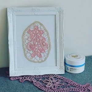 Può includere: Un'immagine incorniciata bianca con un pizzo all'uncinetto rosa e crema all'interno. Il pizzo ha la forma di un ovale. Un barattolo di crema al latte di capra è seduto su un tavolo di fronte all'immagine.