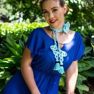 Pu&ograve; includere: Una donna indossa un vestito blu con una collana a forma di farfalla in crochet blu chiaro. La collana ha dei pon pon ed &egrave; un pezzo di grande impatto.
