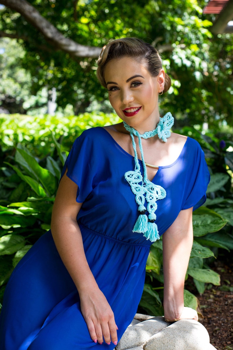 Pu&ograve; includere: Una donna indossa un vestito blu con una collana a forma di farfalla in crochet blu chiaro. La collana ha dei pon pon ed &egrave; un pezzo di grande impatto.