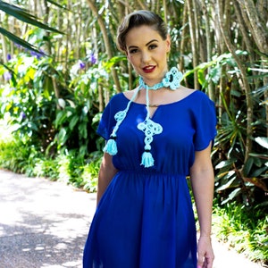 Pu&ograve; includere: Una donna indossa un abito blu reale con una collana di pizzo blu chiaro con nappe. &Egrave; in piedi in un giardino.