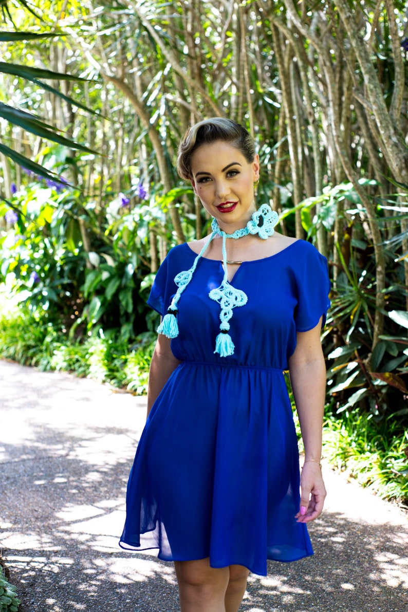 Pu&ograve; includere: Una donna indossa un abito blu reale con una collana di pizzo blu chiaro con nappe. &Egrave; in piedi in un giardino.