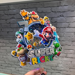 May include: A colorful Super Mario-themed cake topper featuring Mario, Luigi, Bowser, Princess Peach, Yoshi, and other characters. The number 7 is displayed, along with the name "Super Ricky". The topper is made of layered cardstock and has a clear plastic stick.