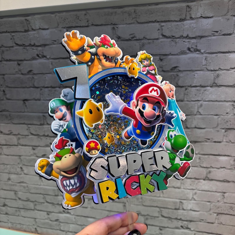 May include: A colorful Super Mario-themed cake topper featuring Mario, Luigi, Bowser, Princess Peach, Yoshi, and other characters. The number 7 is displayed, along with the name "Super Ricky". The topper is made of layered cardstock and has a clear plastic stick.