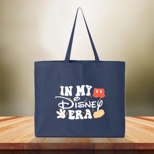 Puede incluir: Bolso tote azul marino con el texto "IN MY Disney ERA" en blanco, con gr&aacute;ficos tem&aacute;ticos de Mickey Mouse. El bolso tiene forma rectangular y dos asas.