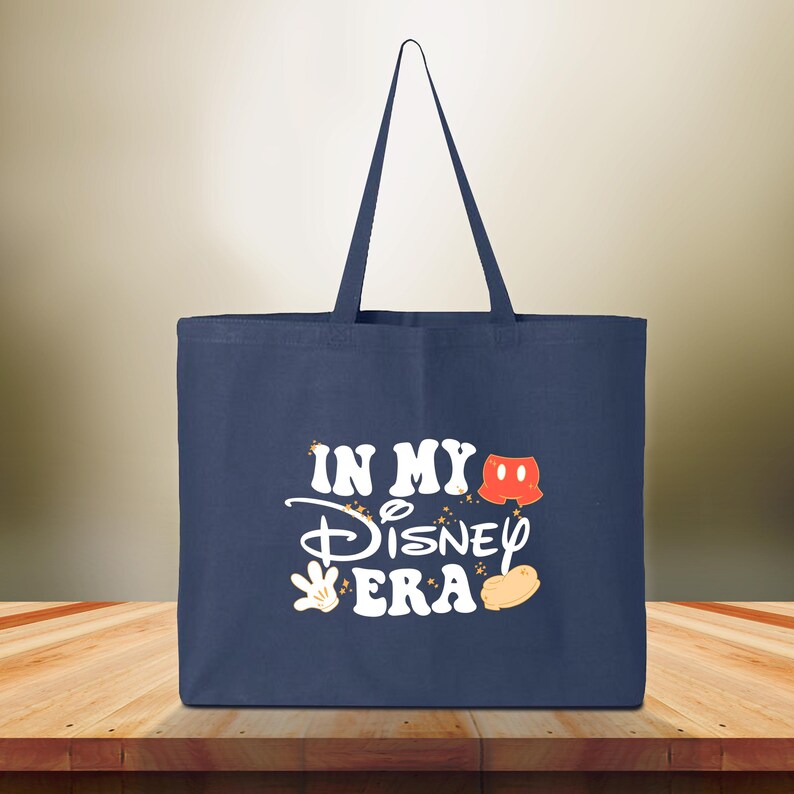 Puede incluir: Bolso tote azul marino con el texto "IN MY Disney ERA" en blanco, con gr&aacute;ficos tem&aacute;ticos de Mickey Mouse. El bolso tiene forma rectangular y dos asas.