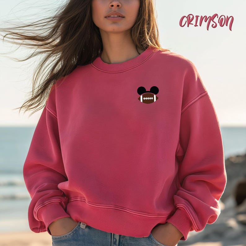 May include: Crimson-colored sweatshirt with a small embroidered design of a football inside black Mickey Mouse ears. The word "Crimson" is written in cursive in the upper right corner. The sweatshirt has a crew neck and long sleeves.