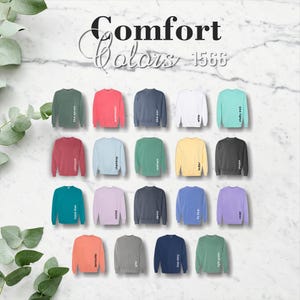 May include: A selection of Comfort Colors 1566 crewneck sweatshirts in various colors, including blue spruce, watermelon, blue jean, white, chalky mint, crimson, chambray, and more. The sweatshirts are displayed on a marble background.
