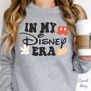 May include: A light gray sweatshirt with the text "IN MY Disney ERA" in black, with Mickey Mouse-themed graphics. The sweatshirt also features a pair of red shorts, a white glove, and a yellow shoe.