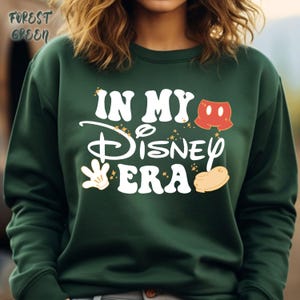 May include: Forest green sweatshirt with the text "IN MY Disney ERA" in white, with Mickey Mouse-themed graphics. The sweatshirt features a crew neck and long sleeves. The text is accompanied by star accents and Mickey Mouse shorts, glove, and shoe graphics.