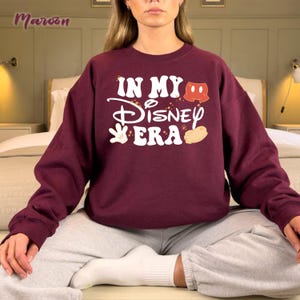 May include: A maroon sweatshirt with the text "IN MY Disney ERA" in white, with Mickey Mouse-themed graphics. The sweatshirt is long-sleeved and has a crew neck. The word "Maroon" is printed in the upper left corner.