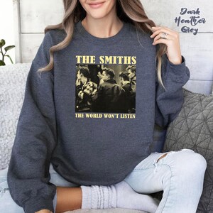 May include: Dark heather grey sweatshirt featuring a sepia-toned image of a band with the text "THE SMITHS" above and "THE WORLD WON'T LISTEN" below. The sweatshirt is a crew neck style.