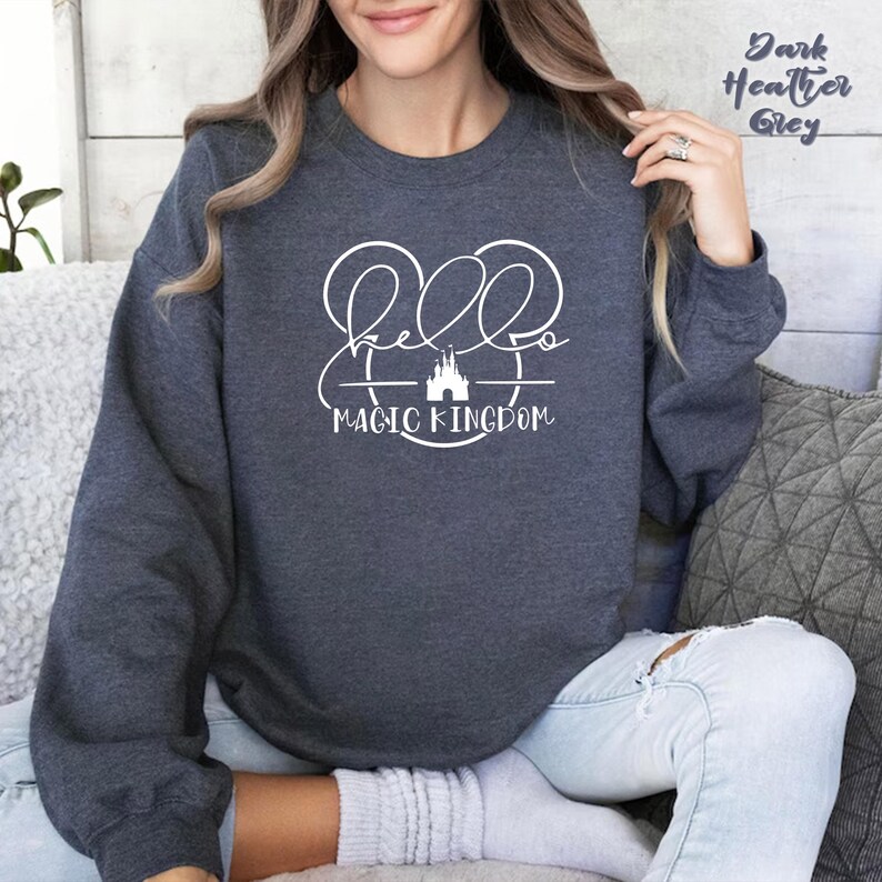 May include: Dark heather grey sweatshirt with the text "Hello Magic Kingdom" in white script and a castle graphic. The sweatshirt has a crew neck and long sleeves. The text "Dark Heather Grey" is in the upper right corner.