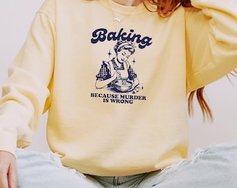 May include: A light yellow sweatshirt with the word "Baking" in navy blue script above a vintage illustration of a woman mixing ingredients. The text "BECAUSE MURDER IS WRONG" is below the illustration.