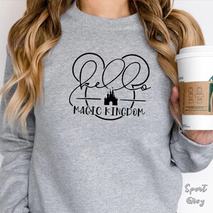 May include: A light gray sweatshirt with the words "Hello Magic Kingdom" in a black script font. The design includes a silhouette of a castle and Mickey Mouse ears. The sweatshirt is labeled "Sport Grey".