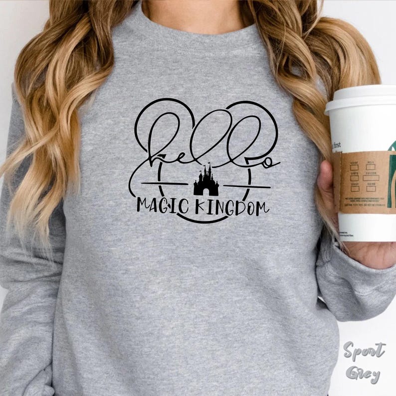 May include: A light gray sweatshirt with the words "Hello Magic Kingdom" in a black script font. The design includes a silhouette of a castle and Mickey Mouse ears. The sweatshirt is labeled "Sport Grey".