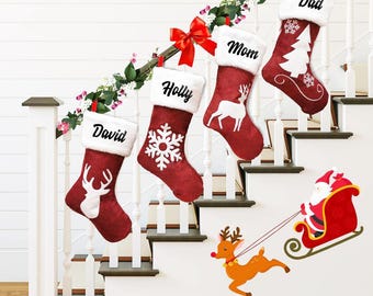 May include: Four red Christmas stockings with white faux fur cuffs hang on a staircase. Each stocking is personalized with a name and features a different holiday design, including a snowflake, a deer, and a Christmas tree. A decorative garland and a Santa and reindeer decal complete the festive scene.