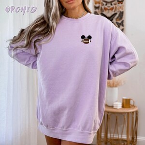 May include: A light purple sweatshirt with a small graphic of a football with Mickey Mouse ears. The word "ORCHID" is printed in a light purple script at the top left. The sweatshirt is a solid color and has long sleeves.