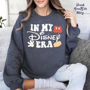 May include: Dark heather grey sweatshirt with the text "IN MY Disney ERA" in white, with Mickey Mouse-themed graphics. The sweatshirt has a crew neck and long sleeves. The text "Dark Heather Grey" is printed in the upper right corner.