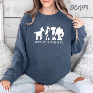 May include: Denim blue sweatshirt with a white graphic of cartoon characters and the text "YOU'VE GOT A FRIEND IN ME". The sweatshirt is a crew neck style and has long sleeves. The word "Denim" is written in cursive at the top right.