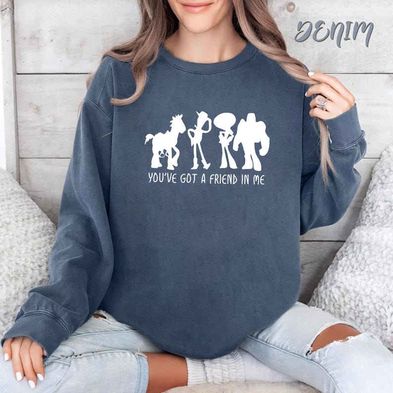 May include: Denim blue sweatshirt with a white graphic of cartoon characters and the text "YOU'VE GOT A FRIEND IN ME". The sweatshirt is a crew neck style and has long sleeves. The word "Denim" is written in cursive at the top right.
