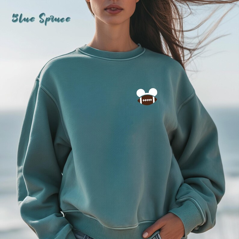 May include: A teal sweatshirt with a white and brown football graphic. The text "Blue Spruce" is written in cursive at the top left. The sweatshirt has a crew neck and long sleeves.