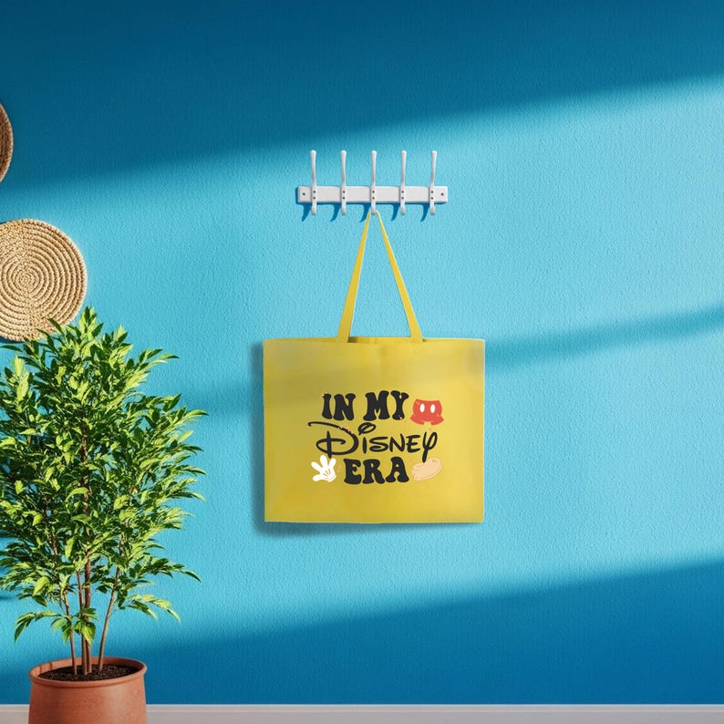 Puede incluir: Un bolso tote amarillo con el texto "IN MY Disney ERA" en escritura negra y un gr&aacute;fico de Mickey Mouse. La bolsa cuelga de un perchero blanco en una pared azul. Tambi&eacute;n se ven una maceta y adornos de pared.