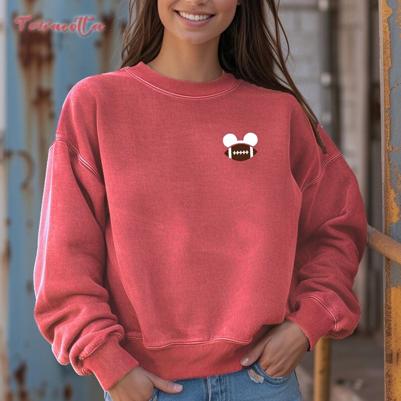 May include: Red-orange sweatshirt with a small graphic of a football and Mickey Mouse ears. The sweatshirt has long sleeves and a crew neck. The graphic is on the upper left chest area. The sweatshirt is made of a soft material.