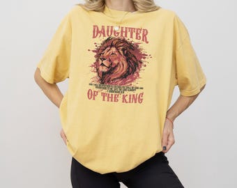 May include: Mustard yellow t-shirt with the text "DAUGHTER OF THE KING" and a lion graphic. The lion graphic is in shades of brown and red. The shirt also has a quote from 2 Corinthians 6:18.
